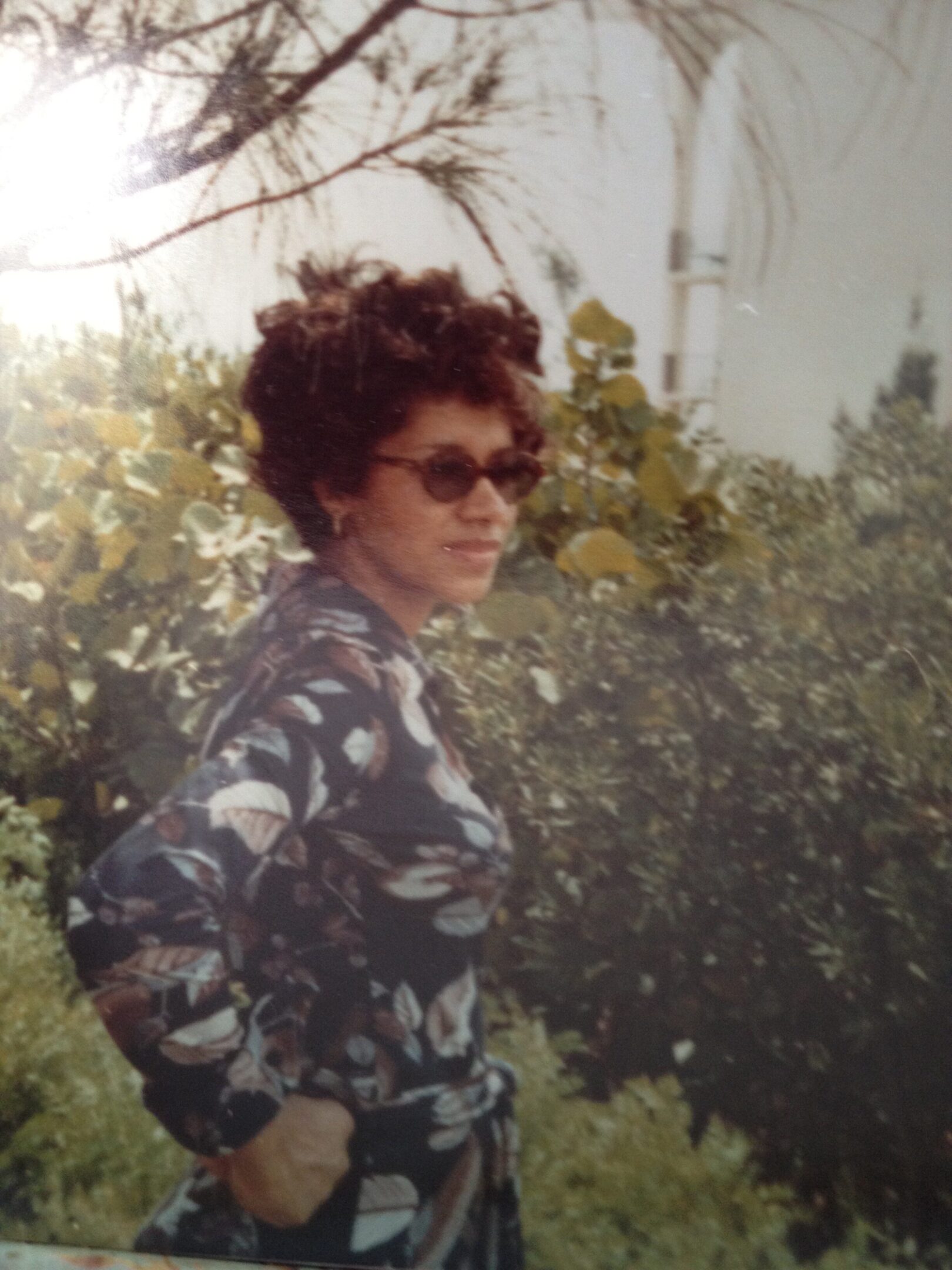 A woman in sunglasses standing next to bushes.